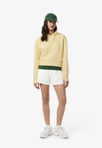 Gelbe, cropped Kapuzenjacke, grüne Schicht darunter, weiße Shorts und weiße Sneaker. Grüner Hut. Weiches Material, lässige Passform. Minimale Details.