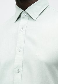 Šviesiai mėtinės spalvos marškiniai su mygtukais, pagaminti iš tekstūruotos medžiagos, su klasikinio stiliaus apykakle ir mažyčiu siuvinėtu logotipu ant krūtinės.