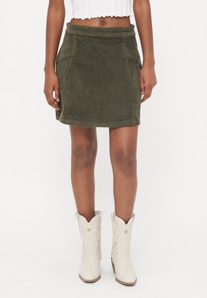 Femme portant une mini-jupe en velours côtelé vert olive avec poches avant, un top court blanc et des bottes de cowboy blanches brodées.