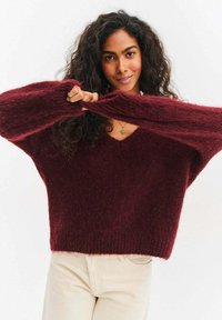 Bordowy sweter dziany z dekoltem w serek, o oversize'owym kroju i długich rękawach, wykonany z miękkiej, teksturowanej tkaniny. Minimalistyczny design bez wzorów.