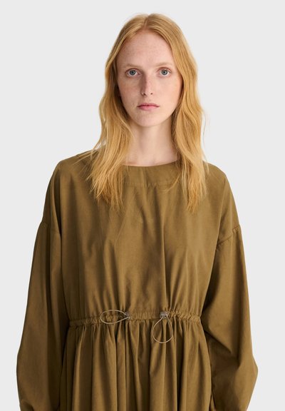 Femme aux longs cheveux roux portant une robe ample vert olive avec des cordons ajustables à la taille, debout devant un fond clair uni.