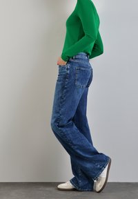 Blå denimjeans med trumpetben, med medelhög midja, framfickor och bakfickor, ihopparat med en figurnära grön långärmad topp och vita sneakers.