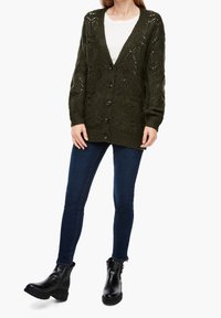 Dunkelgrüne Strickjacke mit tiefem V-Ausschnitt, mit Knopfleiste und kunstvollen Spitzenmustern, getragen über einem weißen Hemd und blauen Jeans.