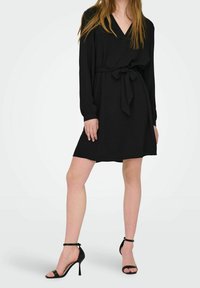 Black long-sleeve dress with a V-neck, waist tie detail, and a loose fit. Paired with black strappy high-heeled sandals.