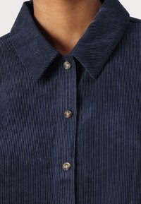 Navy blue corduroy shirt with a collar, featuring vertical ribbed texture and metallic buttons along the front. Close-up of upper section.
