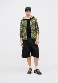 Olivza zaļās krāsas kamuflāžas jaka ar rāvējslēdzēju, melni šorti, melni sporta apavi un melna cepure, ar brīvu piegriezumu un vairākām kabatām.