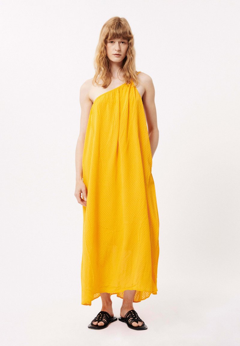 Gelbes, ärmelloses Maxikleid aus leichtem Stoff, mit einem strukturierten Muster und asymmetrischem Saum, kombiniert mit schwarzen Sandalen.