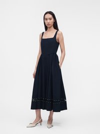 Femme vêtue d'une robe midi noire sans manches avec un décolleté carré et un détail à œillets près de l'ourlet, associée à des talons slingback clairs.