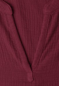 Gros plan d’un col en V profond sur un tissu texturé et doux de couleur bordeaux foncé, montrant les détails des coutures.