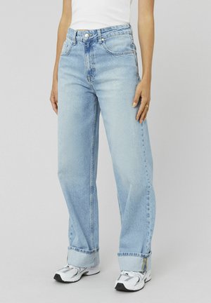 Personne portant un jean large bleu clair avec les ourlets retroussés et des baskets blanches, sur un fond gris uni.