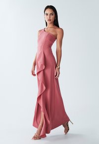 Abito lungo in tessuto rosa tenue, con design a spalla unica, dettaglio laterale con volant, corpetto aderente e spacco laterale. Sandali con tacco dorati e cinturini.