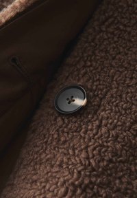 Tissu marron avec une surface texturée et duveteuse, mettant en valeur un bouton noir à quatre trous. L'arrière-plan présente un matériau lisse.