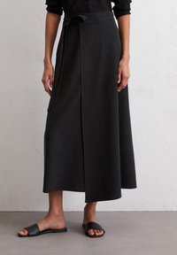 Gonna a portafoglio nera, lunga fino al ginocchio, con vita legata, realizzata in tessuto morbido. Presenta due tasche frontali e una superficie liscia. Abbinata a sandali neri a scivolo.