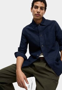 Granatowa koszula zapinana na guziki z kołnierzykiem, dwiema kieszeniami na piersi i podwiniętymi rękawami; noszona z oliwkowymi spodniami.