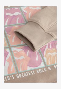 El puño de un suéter beige descansa sobre un estampado gráfico que presenta labios coloridos en tonos rosa y durazno, con detalles en blanco y texto en negrita debajo.