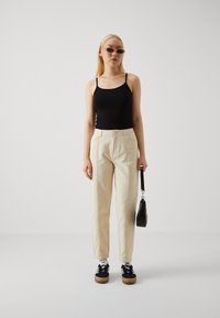 Svart ärmlös topp, krämfärgade byxor med veck, svarta sneakers med gummisulor och en liten svart handväska. Modellen har rakt blont hår.