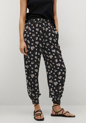 Woman wearing black floral-print loose pants with ankle cuffs, paired with black strappy flat sandals and a sleeveless black top.