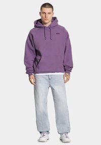 Fioletowa bluza z kapturem ze sznurkiem i kieszenią z przodu, zestawiona z jasnoniebieskimi luźnymi dżinsami i białymi trampkami. Proste czarne logo na bluzie.