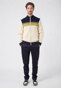 Tröja i kremfärg med marinblå krage, med horisontella ränder i grått och grönt. Slimfit marinblå byxor och svarta skor med vita detaljer.