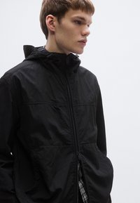 Must kerge kapuutsiga jakk ees tõmblukuga ja küljetaskutega. Iseloomustab mati tekstuur ja minimalistlik disain.