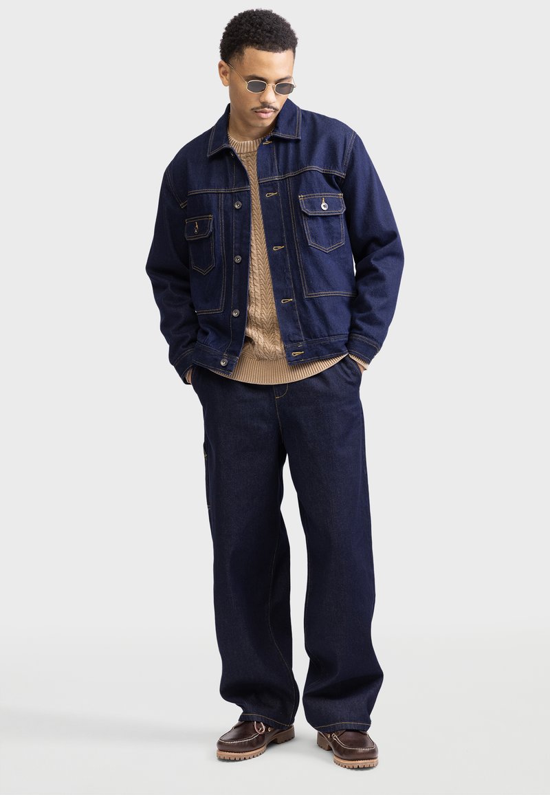 Marineblaue Jeansjacke mit zwei Fronttaschen, beige Kabelstrickpullover und weit geschnittene Hose, kombiniert mit braunen Schuhen und runden Sonnenbrillen.