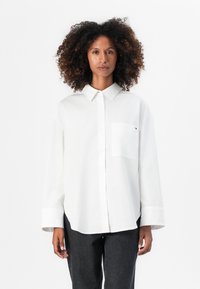 Camisa blanca de manga larga con botones en un tejido suave, con un bolsillo en el pecho y un corte relajado, que presenta un collar puntiagudo y mangas abotonadas.