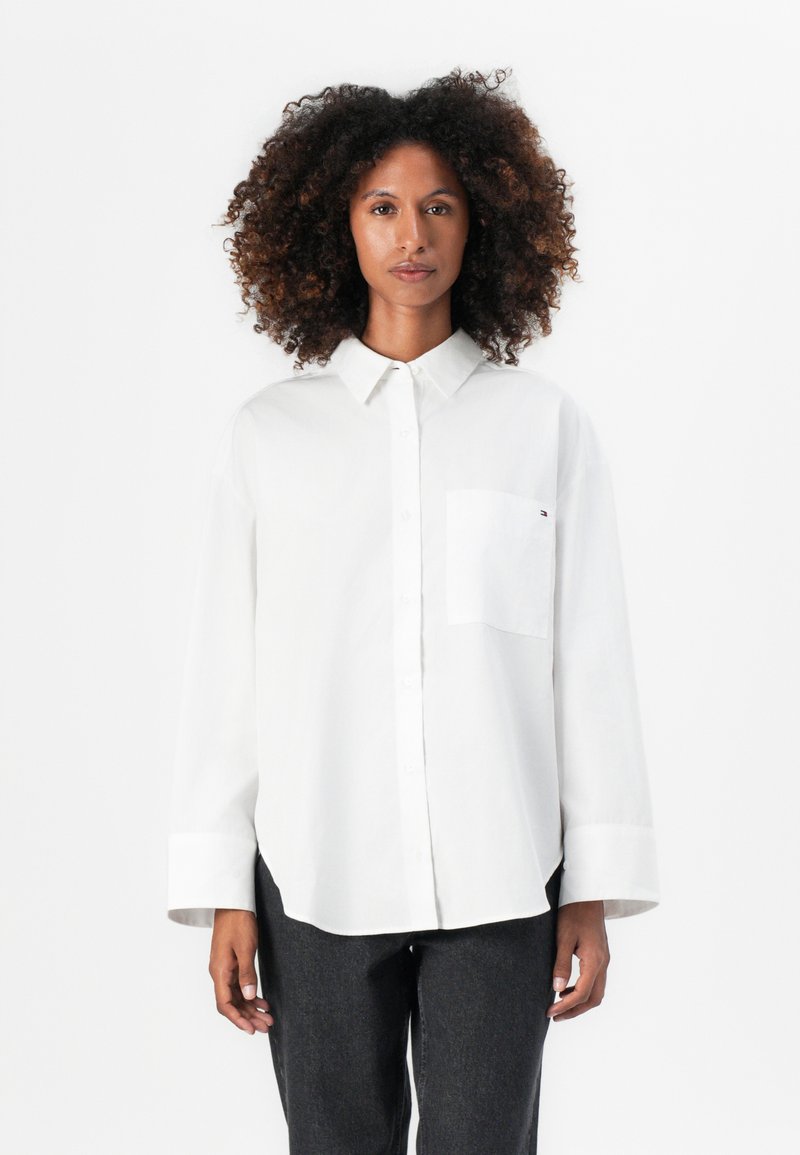 Camisa blanca de manga larga con botones en un tejido suave, con un bolsillo en el pecho y un corte relajado, que presenta un collar puntiagudo y mangas abotonadas.