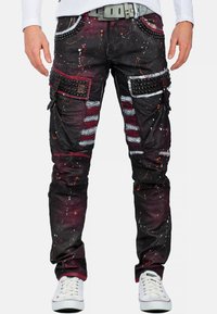 Zwarte cargo broeken met meerkleurige verfspray, textuurstof, versieringen op de zakken en ribbelpatronen. Gecombineerd met witte sneakers.