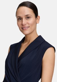 Robe bleu marine avec un design drapé et enroulé, sans manches. La texture du tissu lisse et le détail subtil du col renforcent sa structure.