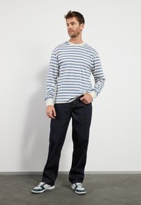 Langermet stripet skjorte i blått og hvitt, kombinert med mørke jeans og flerfargede joggesko, plassert mot en enkel bakgrunn.
