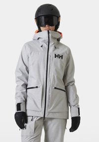 Šedá lyžařská bunda z texturované látky s vysokým límcem, nastavitelnou kapucí, černými akcenty a dvěma předními zipovými kapsami.