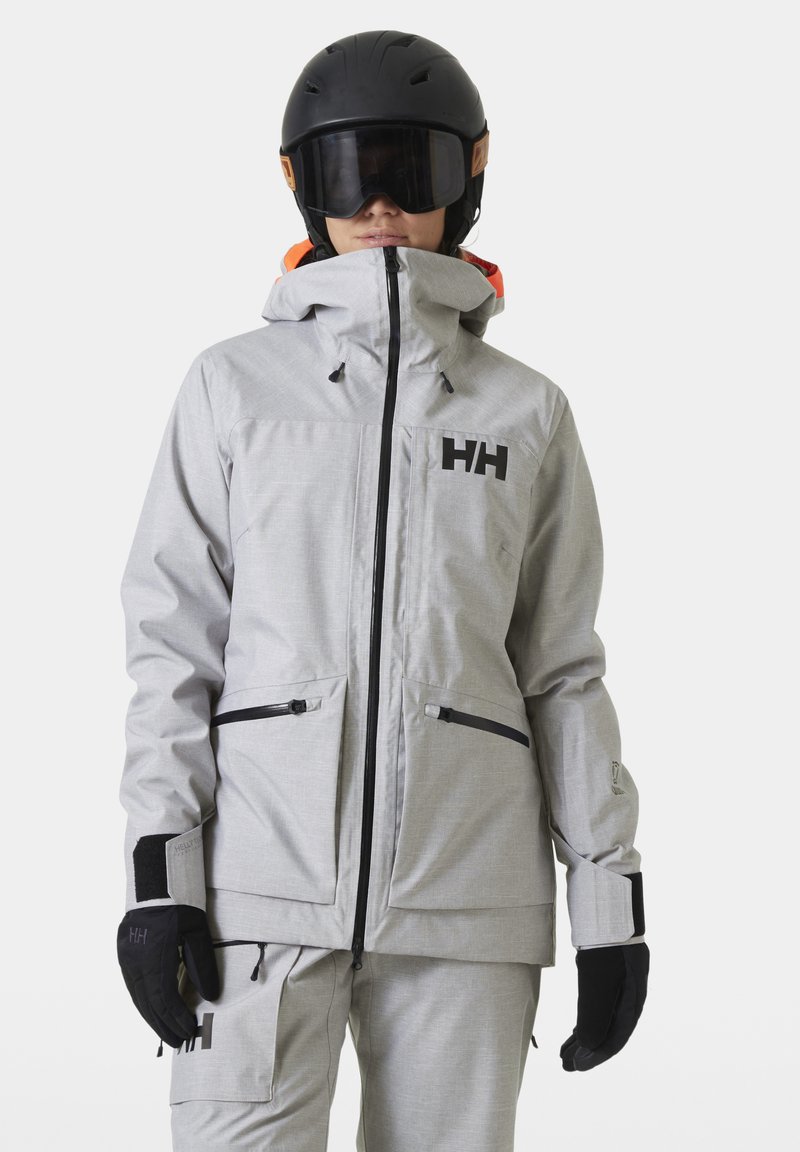 Šedá lyžařská bunda z texturované látky s vysokým límcem, nastavitelnou kapucí, černými akcenty a dvěma předními zipovými kapsami.