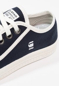 Navy canvas sneaker with white laces, rubber sole, and logo detail on the side. Features rounded toe and cushioned interior.
