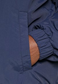 Granatowa kurtka z marszczonymi mankietami, z boczną kieszenią z prostym szwem. Gładki, lekki materiał o delikatnym połysku.