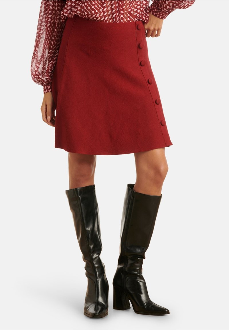 Red wool skirt with a flared shape, side button detailing, and a textured finish, paired with knee-high black leather heeled boots.