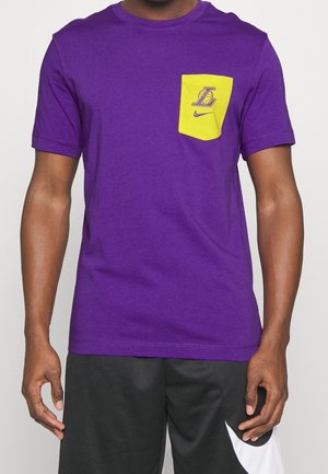 Hombre que lleva una camiseta morada de Los Angeles Lakers con bolsillo amarillo y pantalones cortos negros de Nike con logo blanco de swoosh.