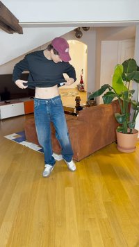 Musta pitkähihaine paita, siniset farkut, hopea urheilukengät; seisoo huoneessa, jossa on ruskea vakosametisohva, vihreä kasvi ja puulattia.