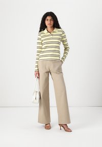 Gestreepte lange mouw polo in geel en bruin, gecombineerd met beige wijde broekspijpen. Model houdt een kleine witte handtas vast en draagt bruine hoge hakken.