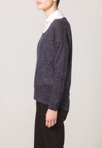 Maglione blu scuro, vestibilità ampia, texture leggera in maglia, design a V, maniche lunghe, indossato su una camicia bianca con colletto, abbinato a pantaloni neri.