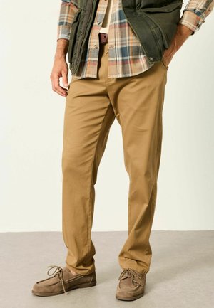 Mann trägt beige Khakihose, braune Wildlederschuhe, ein kariertes Hemd und eine dunkelgrüne Weste, steht mit den Händen nahe den Taschen.