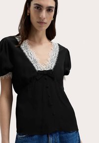 Blouse noire avec un décolleté en V bordé de dentelle blanche, manches bouffantes, accent nœud et fermeture à boutons sur le devant. Tissu doux avec des détails texturés.