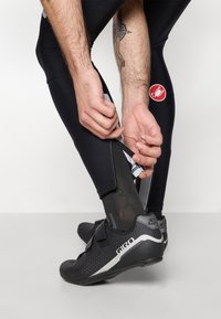 Des collants de cyclisme noirs avec une fermeture éclair à la cheville et un logo rouge sur la cuisse. La personne porte des chaussures de cyclisme noires avec des accents gris.