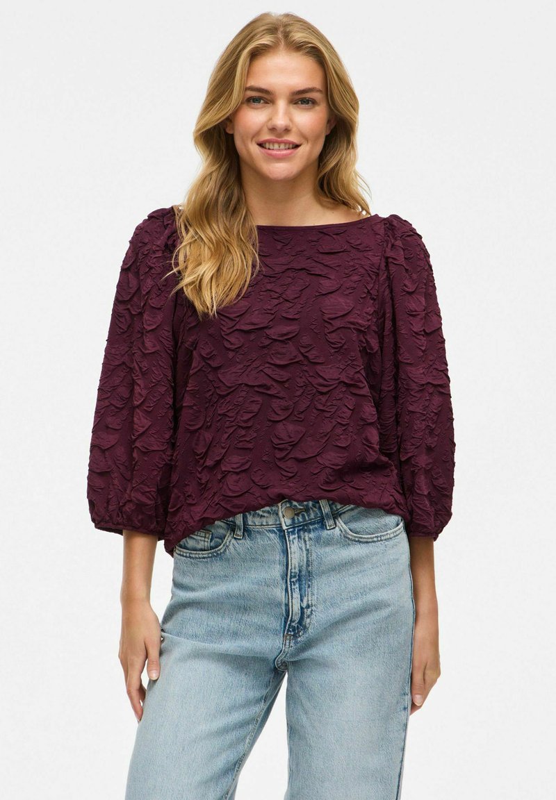 Burgundská blůza s texturovanou tkaninou a nabranými rukávy, kombinovaná se světle modrými džínami. Blůza má široký výstřih a volný střih.