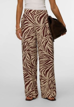 Kvinne iført beige vidbukser med mørkebrunt zebramønster, hvit ermeløs topp, holder en mørkebrun veske og har på seg sandaler.