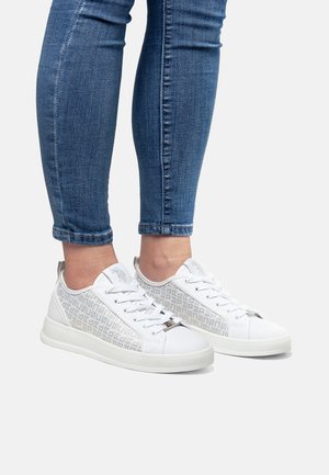 Baskets blanches avec un motif géométrique texturé sur les côtés, lacets blancs et semelle en caoutchouc solide. Un jean avec des ourlets retroussés est porté.