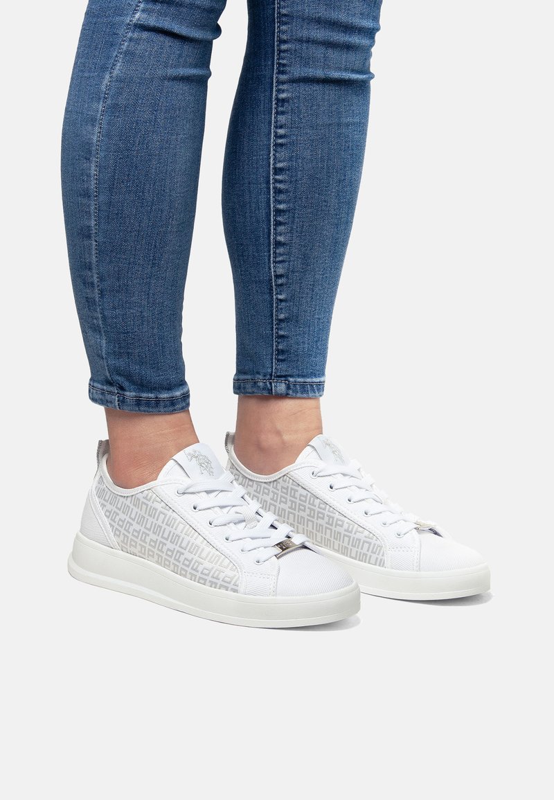 Zapatillas blancas con un patrón geométrico texturizado en los laterales, cordones blancos y una suela de goma sólida. Se llevan vaqueros con dobladillos enrollados.