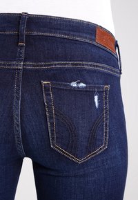 Jeans de mezclilla azul oscuro con un bolsillo trasero desgastado, con ligeras deshilachaduras y costuras doradas contrastantes. Etiqueta de cuero en la cintura.