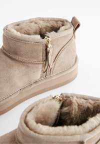 Beige suede ankle boots with faux fur lining, side zip closure, and a textured rubber sole. Features a small heel and stitching details.