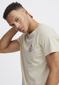 Beige bomulds t-shirt med korte ærmer, prydet med et farverigt broderet grafik af en pære og tekst. Glat tekstur og afslappet pasform.