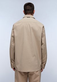 Camisa de manga larga beige hecha de una tela suave, con un cuello puntiagudo, puños con botones y un dobladillo recto. Diseño minimalista sin patrones.